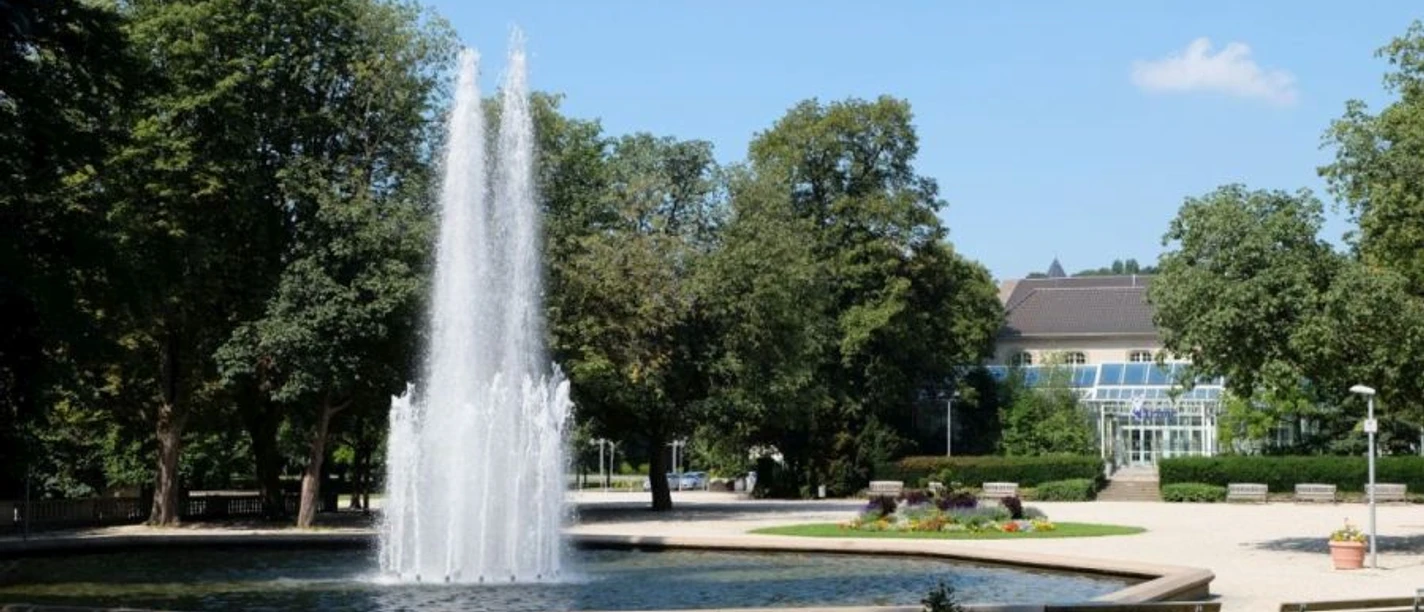 Runder Teich innerhalb einer Parkanlage mit Wasserfontäne in der Mitte.
