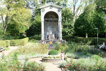 Ein Monument von klassizistischer Architektur umgeben von bunten Blumenbeeten und ruhigen Spazierwegen, auf denen mehrere Personen spazieren.