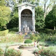 Ein Monument von klassizistischer Architektur umgeben von bunten Blumenbeeten und ruhigen Spazierwegen, auf denen mehrere Personen spazieren.