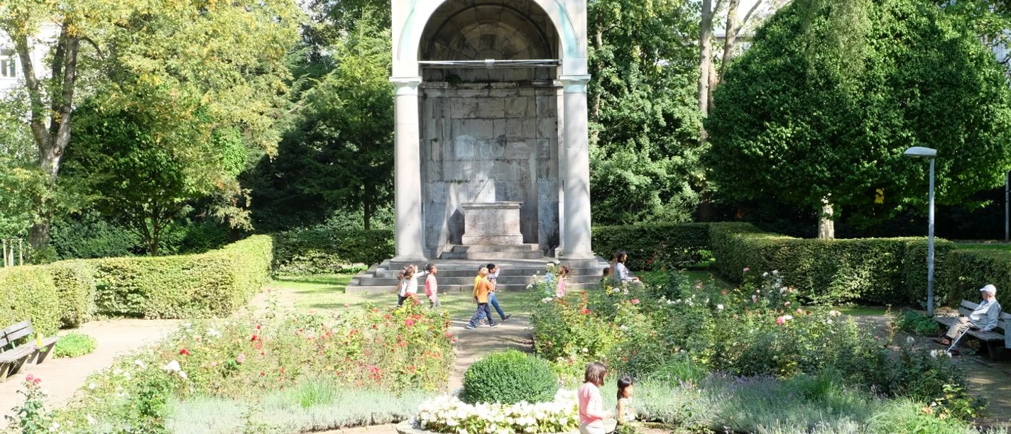 Ein Monument von klassizistischer Architektur umgeben von bunten Blumenbeeten und ruhigen Spazierwegen, auf denen mehrere Personen spazieren.