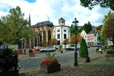 Marktplatz, auf dem geparkte Autos, Laternen, und Blumenkästen stehen, vor einer historischen Kirche.
