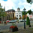 Marktplatz, auf dem geparkte Autos, Laternen, und Blumenkästen stehen, vor einer historischen Kirche.
