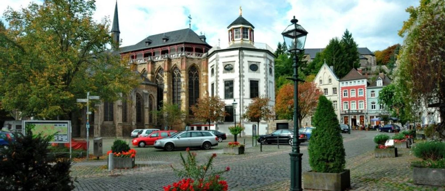 Marktplatz, auf dem geparkte Autos, Laternen, und Blumenkästen stehen, vor einer historischen Kirche.