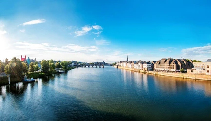 Maastricht Ein breiter Fluss, die Maas, fließt durch eine Stadt. Links grenzen Bäume und Wiesenstreifen an und rechts Häuserreihen.
