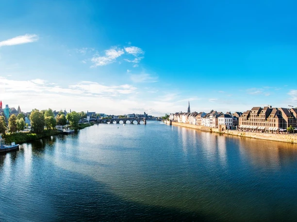 Maastricht Ein breiter Fluss, die Maas, fließt durch eine Stadt. Links grenzen Bäume und Wiesenstreifen an und rechts Häuserreihen.