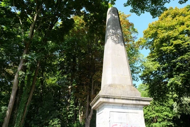 Eine Steinsäule, der Obelisk, steht unter dichten Baumkronen.