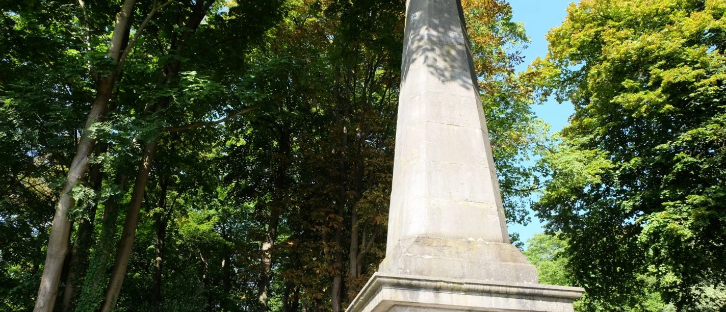 Eine Steinsäule, der Obelisk, steht unter dichten Baumkronen.