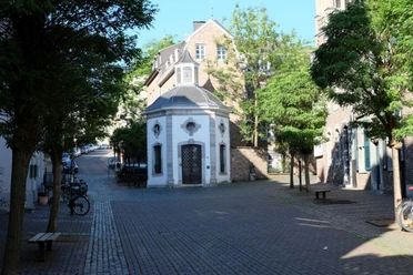 Kleine, achteckige Kapelle mit weißer Fassade und einer markanten Holztür. Sie wird von Bäumen umgeben und steht mitten auf einem gepflasterten Platz, der von weiteren historischen Gebäuden eingerahmt wird.