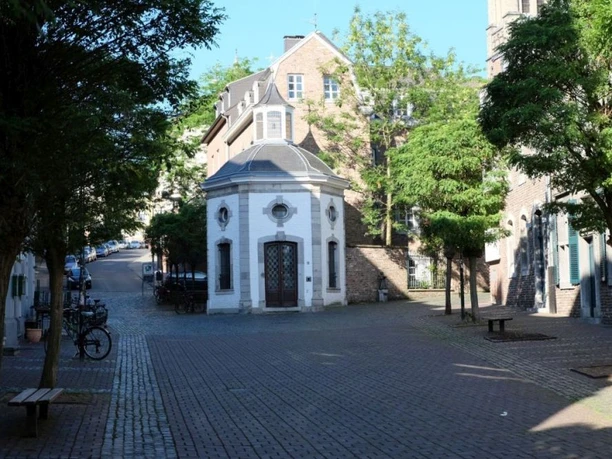 Kleine, achteckige Kapelle mit weißer Fassade und einer markanten Holztür. Sie wird von Bäumen umgeben und steht mitten auf einem gepflasterten Platz, der von weiteren historischen Gebäuden eingerahmt wird.