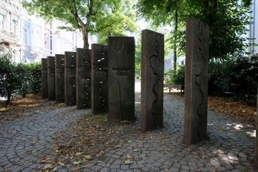 Mahnmal gegen Intoleranz Auf einem kleinen Platz stehen lange Steinblöcke aufrecht wie Dominosteine im Boden. Auf ihnen sind verschiedene geschwungene Linien eingemeißelt.