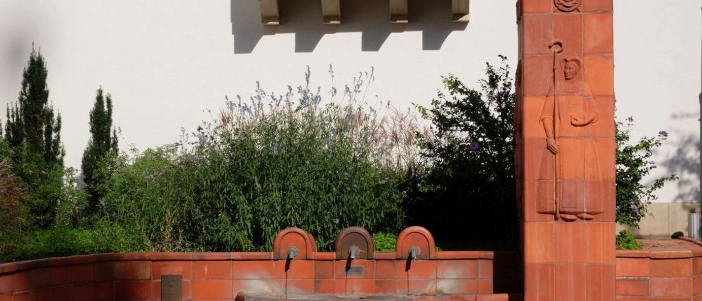 Thermalbrunnen Burtscheid Brunnenbecken eingearbeitet in rotes Gemäuer mit einem Pflanzenbeet im Hintergrund.