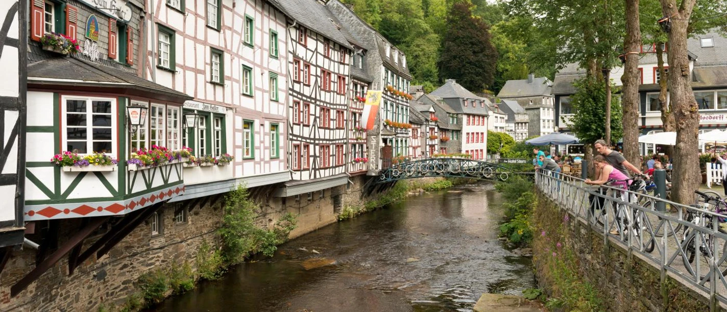 Monschau Traditionelle Fachwerkhäuser entlang eines Flusslaufs mit blühenden Blumenkästen an den Fenstern.