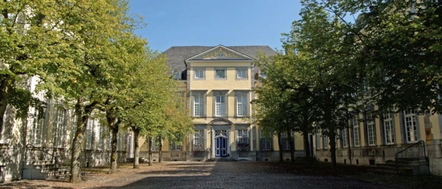 Historisches Gebäude mit gepflastertem Vorplatz zwischen zwei Baumreihen unter blauem Himmel.