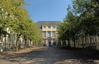 Reichsabtei Kornelimünster Historisches Gebäude mit gepflastertem Vorplatz zwischen zwei Baumreihen unter blauem Himmel.