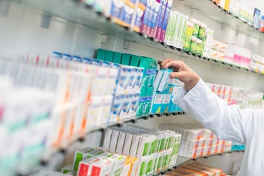 Hirsch Apotheke Arm mit weißem Kittel, der Medikamente aus einem gut sortierten Regal entnimmt, wobei bunte Verpackungen dicht an dicht stehen.