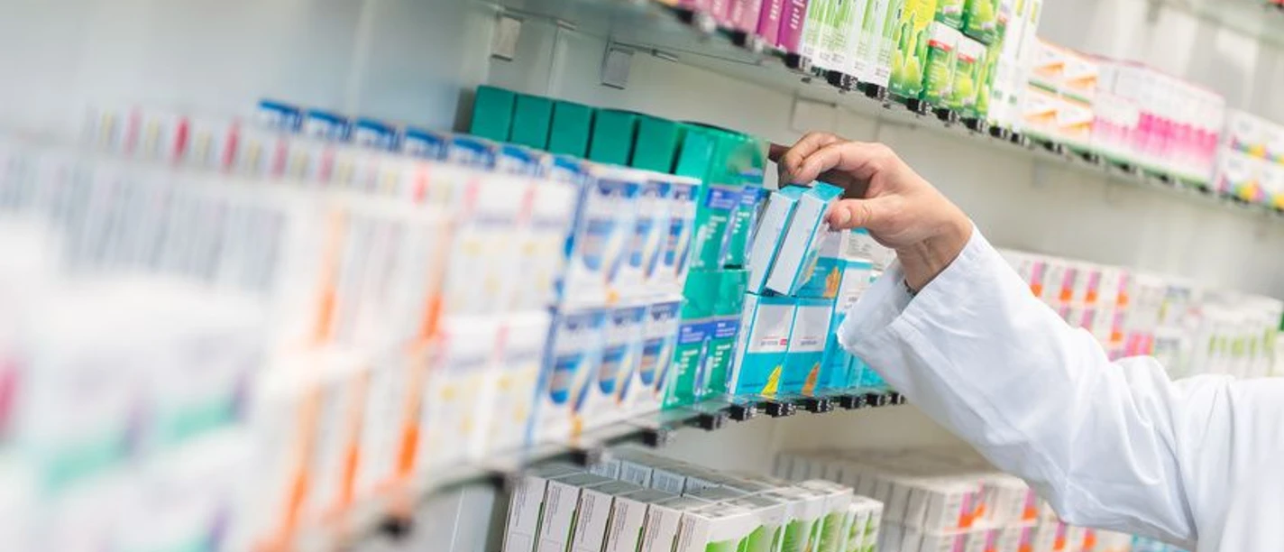 Hirsch Apotheke Arm mit weißem Kittel, der Medikamente aus einem gut sortierten Regal entnimmt, wobei bunte Verpackungen dicht an dicht stehen.