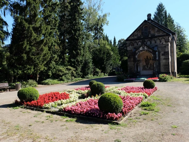 Ostfriedhof Blumenbeet mit rot-weißen Blumen vor einer Friedhofskapelle auf einem kiesigen Weg, umgeben von hohen Bäumen.