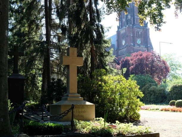 Ostfriedhof Grabstätten umgeben von grünen Bäumen und Sträuchern im Sonnenlicht. Im Hintergrund steht eine Kirche.