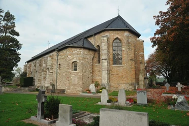 Kirche aus hellbraunen Steinen vor einer Grabesstätte auf einer Wiese, umgeben von herbstlichen Bäumen..