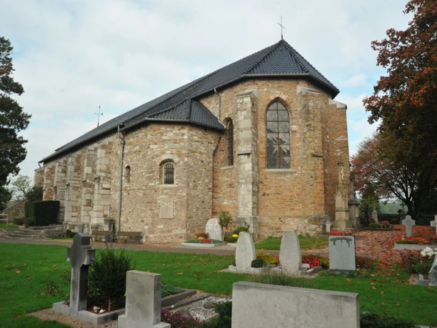 St. Stephanus Kirche aus hellbraunen Steinen vor einer Grabesstätte auf einer Wiese, umgeben von herbstlichen Bäumen..