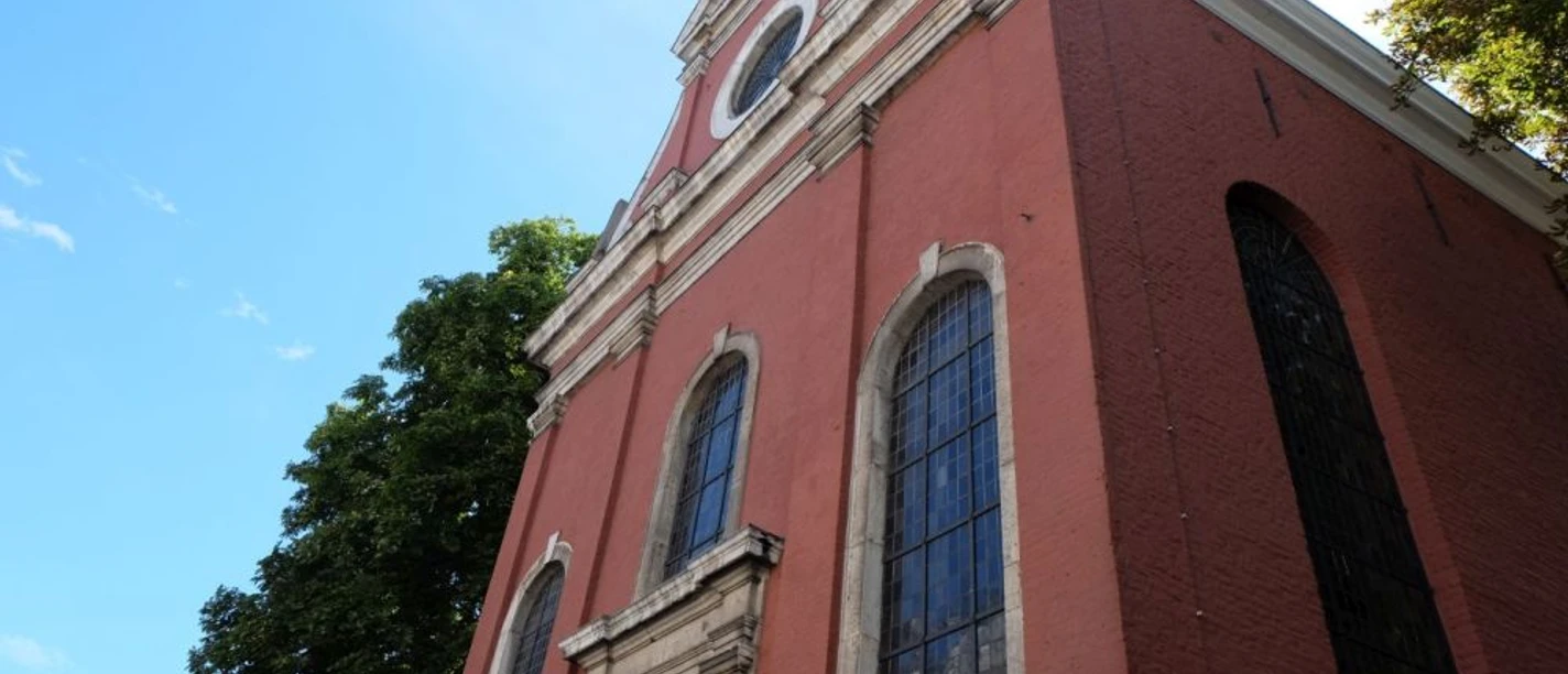 Kirche mit roten Backsteinwänden und großen, hohen Fenstern. Verzierungen sind um die Fenster und an der Fassade. Die Kirche erhebt sich unter einem blauen Himmel, umgeben von Bäumen.