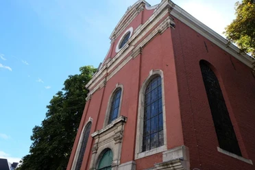 Theresienkirche Kirche mit roten Backsteinwänden und großen, hohen Fenstern. Verzierungen sind um die Fenster und an der Fassade. Die Kirche erhebt sich unter einem blauen Himmel, umgeben von Bäumen.