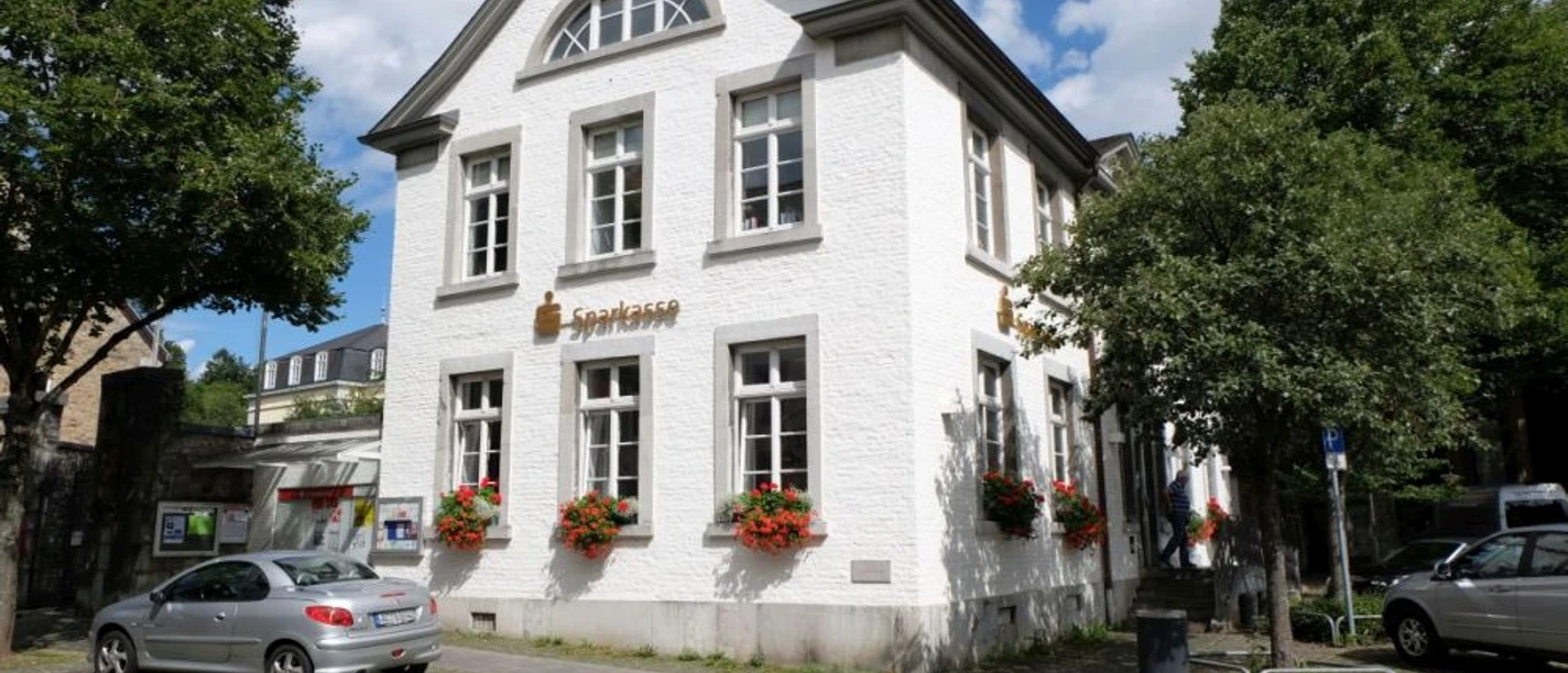 Kornelimünster Weißes, zweistöckiges Gebäude mit dekorativen Blumen, umrahmt von alten Bäumen.