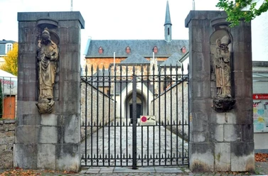Steinerne Patrone Eingangstor zwischen zwei steinernen Heiligenfiguren. Im Hintergrund führt ein Weg zu einem Torbogen.