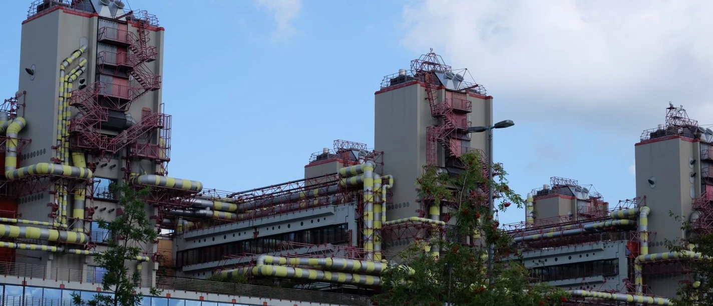 Längliches, industrielles Gebäude mit Glasfront und drei Türmen, an denen rote Treppen und gelbe Rohre verlaufen.