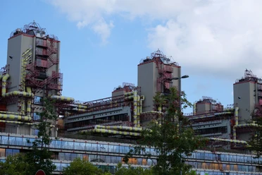 Uniklinik RWTH Aachen Längliches, industrielles Gebäude mit Glasfront und drei Türmen, an denen rote Treppen und gelbe Rohre verlaufen.