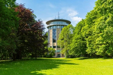 Hinter einer grünen Wiese mit Bäumen an den Seiten steht ein breiter Turm mit gläserner Aussichtsplattform im oberen Geschoss unter blauem Himmel.