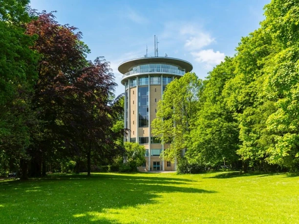 Wasserturm Lousberg Hinter einer grünen Wiese mit Bäumen an den Seiten steht ein breiter Turm mit gläserner Aussichtsplattform im oberen Geschoss unter blauem Himmel.