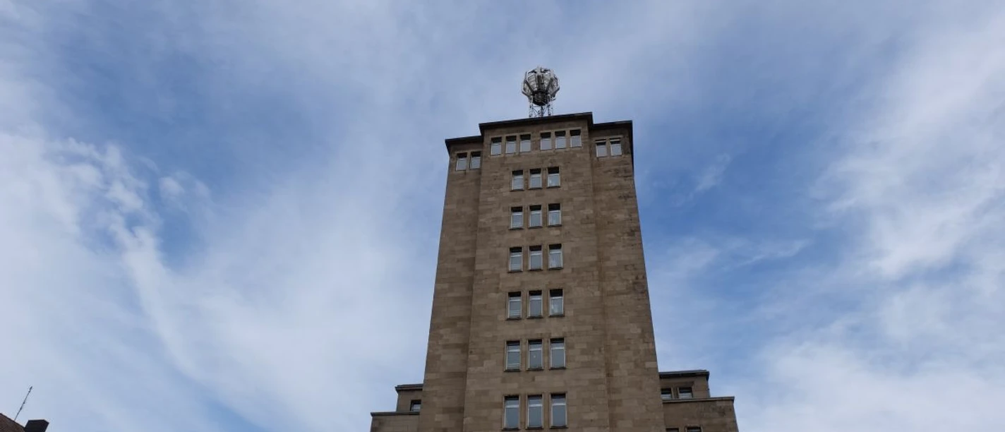 Wettersäule Auf einem hohen, turmähnlichen Gebäude mit Fenstern steht eine Wetterstation mit runder Kuppel unter leicht bewölktem Himmel..