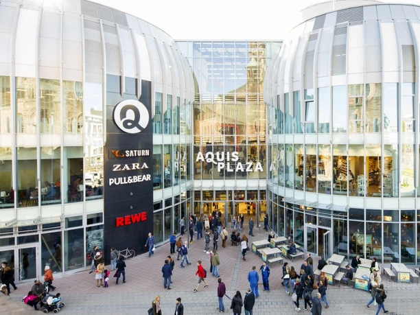 Modernes Einkaufszentrum mit Glasfassade und vielen Besucher*innen. Verschiedene Geschäfte und bekannte Marken sind im Zentrum zu sehen, während zahlreiche Menschen den Haupteingang passieren. Das Design der Gebäude umfasst abgerundete Architekturelemente, die das Licht reflektieren.