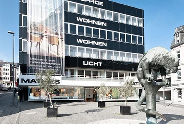 Mathes Modernes, mehrstöckiges Gebäude mit der Skulptur des Bahkauvs im Vordergrund. An den einzelnen Etagen des Gebäudes steht "LICHT", darüber "WOHNEN", darüber "WOHNEN" und ganz oben "OFFICE".