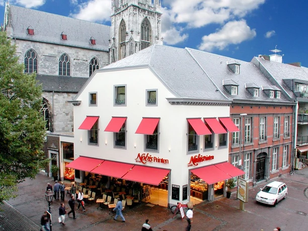 Weißes Eckgebäude mit roten Markisen über den Fenstern und der Aufschrift "Nobis Printen". Unten auf der Straße stehen Stühle und mehrere Personen gehen dort entlang. Im Hintergrund steht eine Kirche.