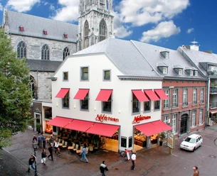 Nobis Printen Weißes Eckgebäude mit roten Markisen über den Fenstern und der Aufschrift "Nobis Printen". Unten auf der Straße stehen Stühle und mehrere Personen gehen dort entlang. Im Hintergrund steht eine Kirche.