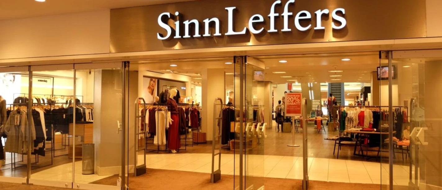 Eingang des Modegeschäfts "SinnLeffers" mit Glastüren. Über dem Eingang steht der Schriftzug "SinnLeffers". Hinter den Glastüren wird Kleidung in Regalen und an Kleiderständern präsentiert.