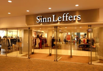 Sinn&Leffers Eingang des Modegeschäfts "SinnLeffers" mit Glastüren. Über dem Eingang steht der Schriftzug "SinnLeffers". Hinter den Glastüren wird Kleidung in Regalen und an Kleiderständern präsentiert.