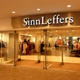 Sinn&Leffers Eingang des Modegeschäfts "SinnLeffers" mit Glastüren. Über dem Eingang steht der Schriftzug "SinnLeffers". Hinter den Glastüren wird Kleidung in Regalen und an Kleiderständern präsentiert.