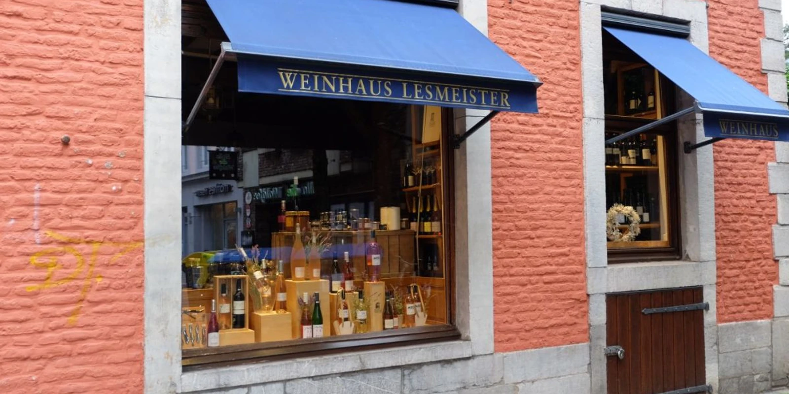 Weinhaus Lesmeister Außenansicht eines Geschäfts mit roter Fassade und blauer Markise über dem Schaufenster, auf dem "Weinhaus Lesmeister" steht.