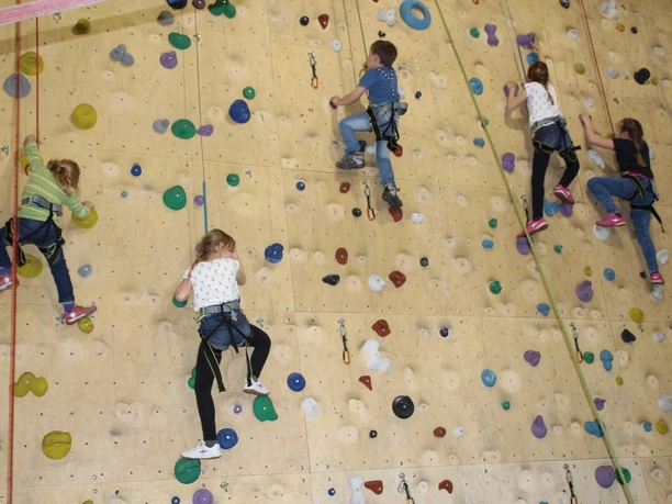 Kletterhalle Abenteuerzeit Kinder klettern gesichert an farbigen Griffen einer hohen Indoor-Kletterwand in der Halle.