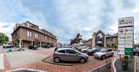 Modehaus Schulte Einkaufszentrum mit Modehaus, Glasfassade und Parkplatz mit mehreren Autos bei bewölktem Himmel.