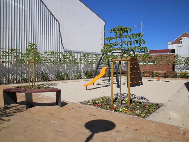 Spielplatz Lange Straße Moderner Spielplatz mit Rutsche, Kletterwand und Sandfläche zwischen Gebäuden unter blauem Himmel.