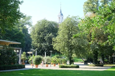 Park mit der Außenterrasse eines Lokals und einer Kirchturmspitze hinter Baumkronen.