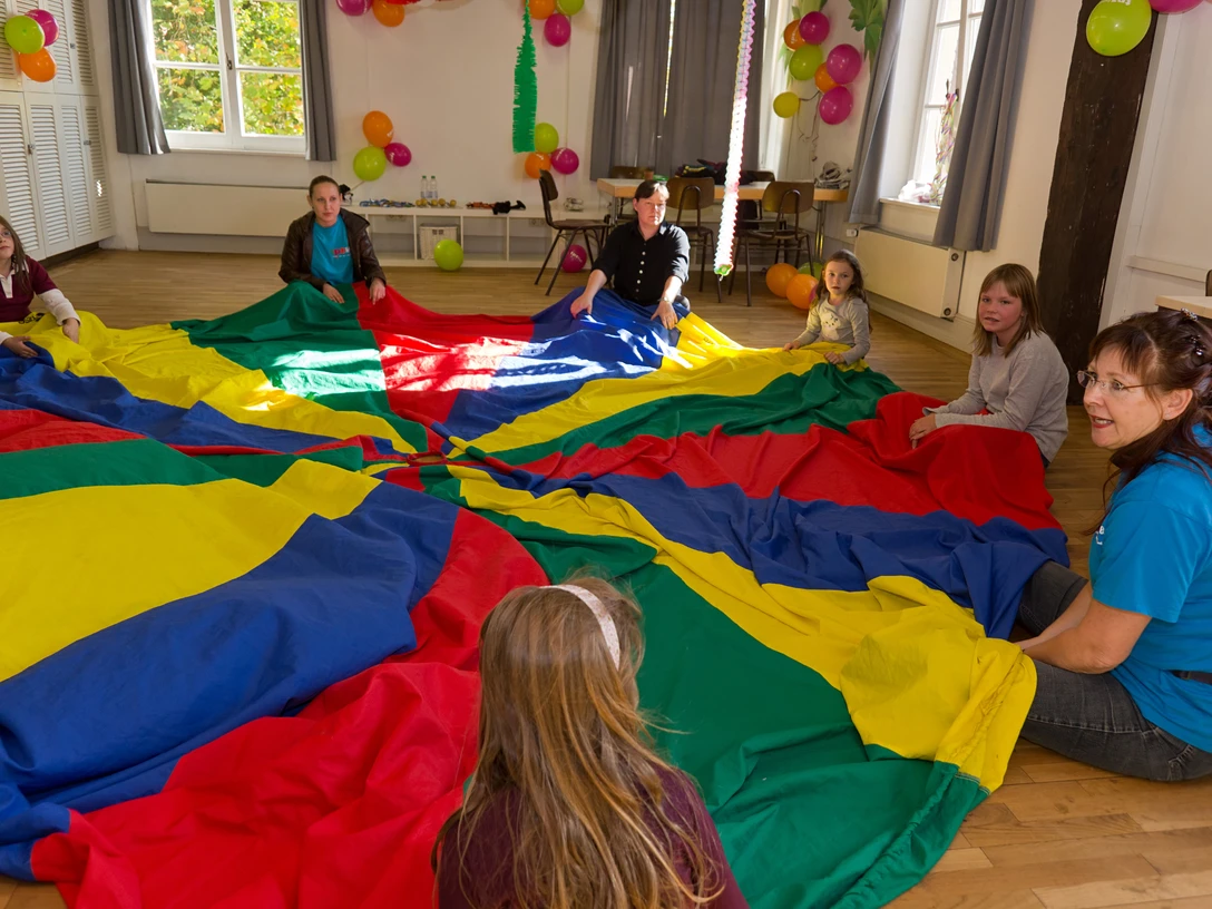 Kinder und Erwachsene sitzen im Kreis und spielen gemeinsam mit einem großen bunten Schwungtuch.