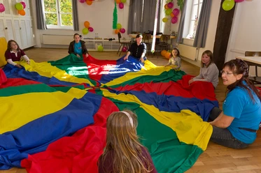 Erlebnishaus im TPZ Lingen Kinder und Erwachsene sitzen im Kreis und spielen gemeinsam mit einem großen bunten Schwungtuch.