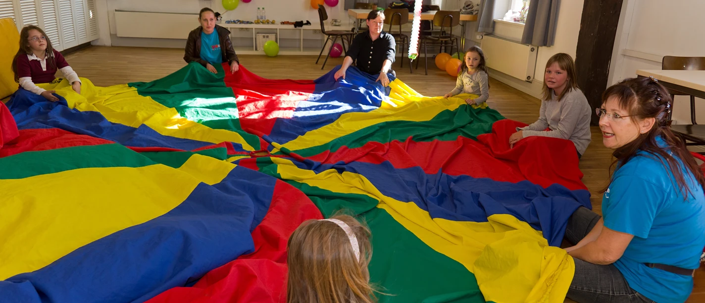 Erlebnishaus im TPZ Lingen Kinder und Erwachsene sitzen im Kreis und spielen gemeinsam mit einem großen bunten Schwungtuch.