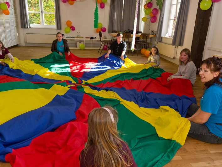 Erlebnishaus im TPZ Lingen Kinder und Erwachsene sitzen im Kreis und spielen gemeinsam mit einem großen bunten Schwungtuch.