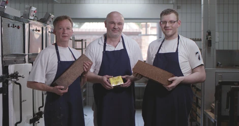 Vollkornbäckerei Ahaus Drei Bäcker in blauen Schürzen stehen in einer Backstube und halten frisch gebackenes Vollkornbrot.
